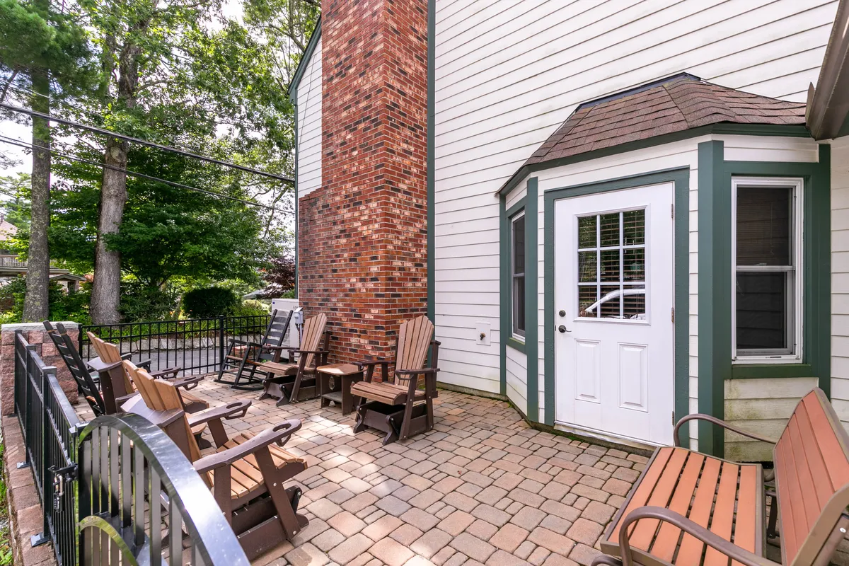 This exterior scene features a brick chimney, wooden chairs, and a white door. The setup invites relaxing with morning coffee or reading outside, with space for socializing and enjoying lovely weather.