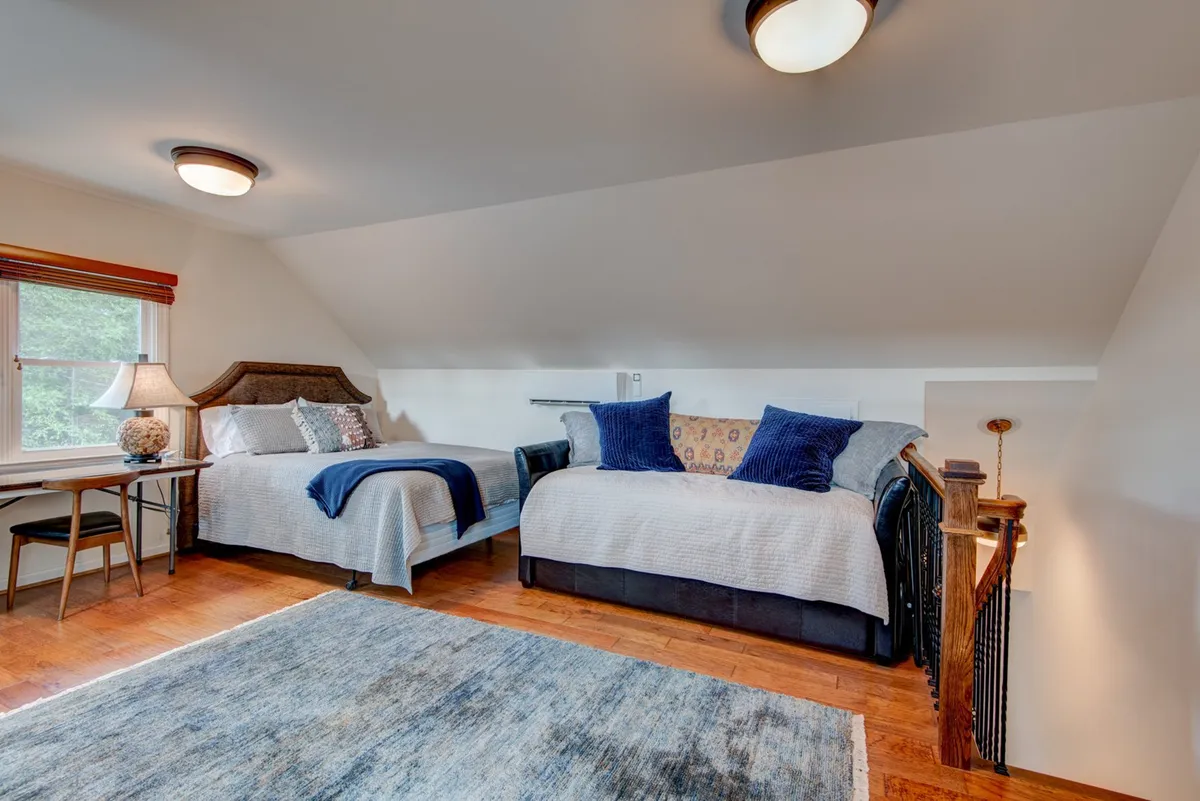 A tranquil bedroom with two beds adorned with decorative pillows, a stylish area rug, and a desk with a lamp by the window. The space feels airy and flexible, suited for families or friends sharing the room.