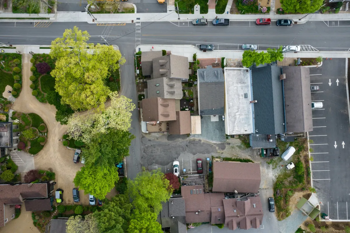 A tranquil scene featuring residential buildings nestled under a leafy tree canopy, with a parking lot close by. The layout suggests an inviting and walkable environment, with easy access for strolling to local shops and community events.