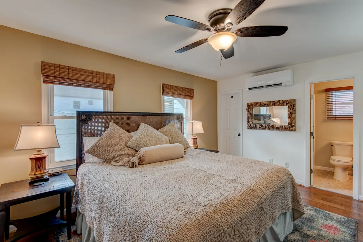 A serene bedroom featuring a large bed dressed in decorative pillows, flanked by two table lamps, with a window letting in natural light. The room is styled with soothing colors, creating an inviting, restful ambiance ideal for unwinding.
