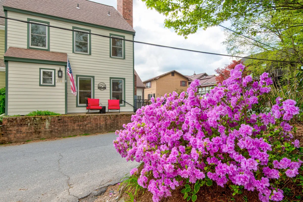 The image displays a cheerful front porch adorned with vibrant pink flowers, red outdoor chairs, an American flag, and the welcoming facade of the home. The blooming flowers and seating area suggest a perfect spot for enjoying the outdoors in spring and summer.