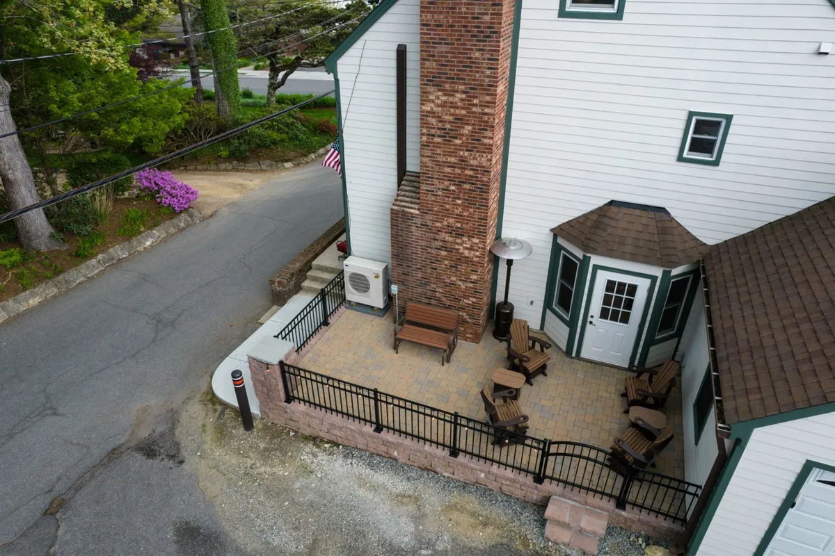 A cozy fenced patio space with comfortable outdoor seating arranged around a small table, framed by landscaped greenery and featuring a prominent brick chimney. The patio is enclosed, offering a sense of privacy while still feeling connected to the outdoor environment.