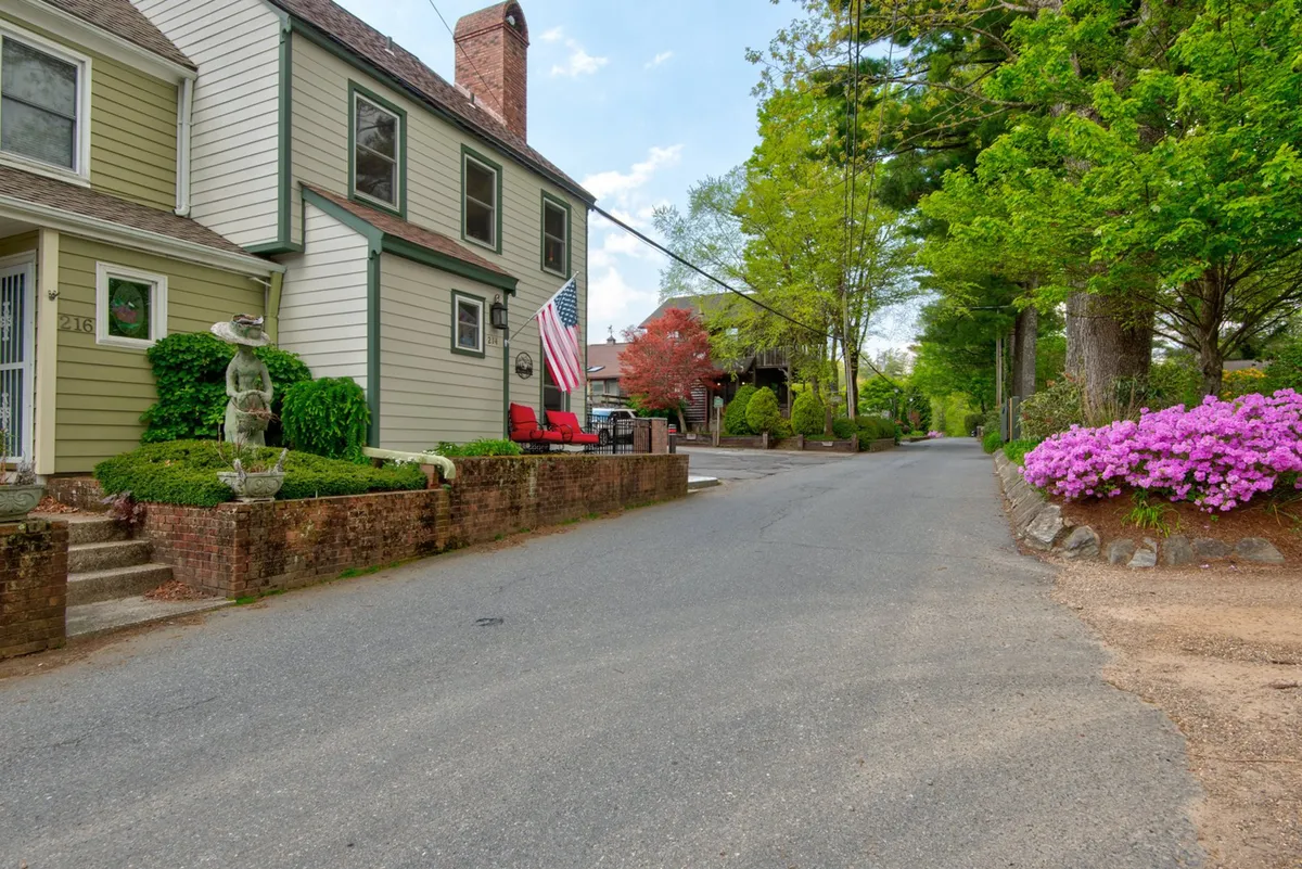 The inviting facade of a home adorned with an American flag, surrounded by vibrant flower beds and a stone wall. Red chairs on the porch beckon, offering a comfortable spot to relax and watch the neighborhood energy unfold. The landscaping and porch create a welcoming transition between home and the lively street beyond.