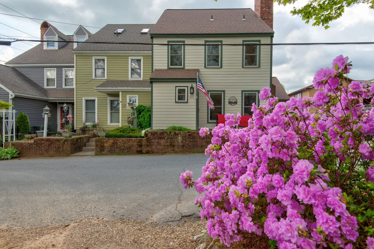 The image showcases the picturesque exterior of The Wander Inn condo, framed by vibrant flowering azaleas, a welcoming front porch adorned with an American flag, and the inviting façade of the home nestled in a friendly, cheerful neighborhood setting.