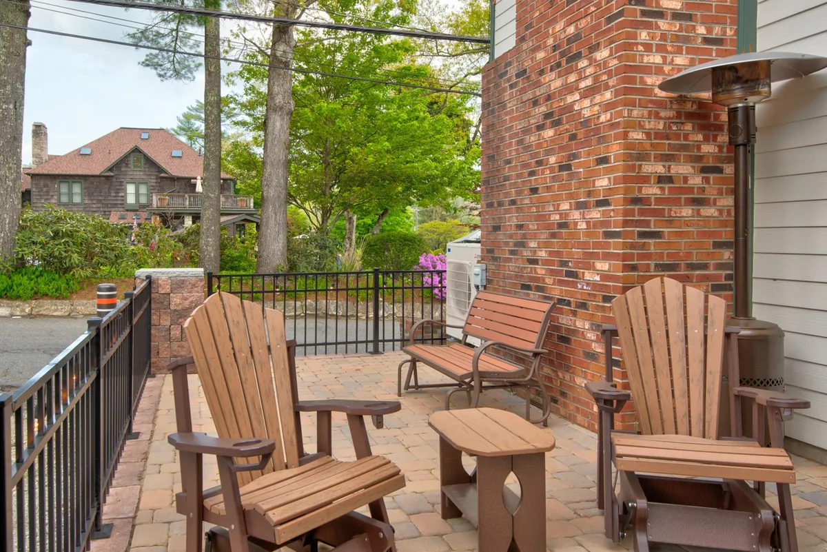 A fenced outdoor seating area featuring wooden chairs and a small table, bordered by a brick wall and colorful seasonal flowers. The enclosure provides privacy while letting in plenty of sunlight, perfect for relaxing or socializing.