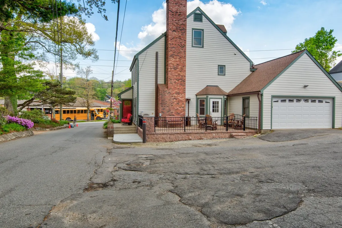 A charming exterior view of The Wander Inn, featuring a welcoming front porch with seating, vibrant flowering bushes, and a classic brick chimney. The scene feels intimate and inviting, suggesting a place where visitors can unwind after a night out.