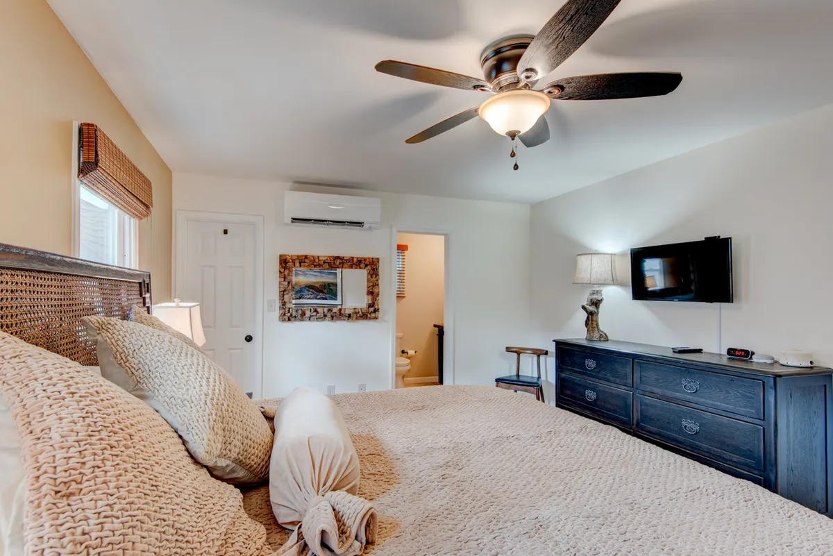 A comfortable bedroom with a bed featuring textured bedding, a ceiling fan overhead, a television, and a nightstand with a lamp. The window allows sunlight to illuminate the room, enhancing its cozy, hotel-like feel.