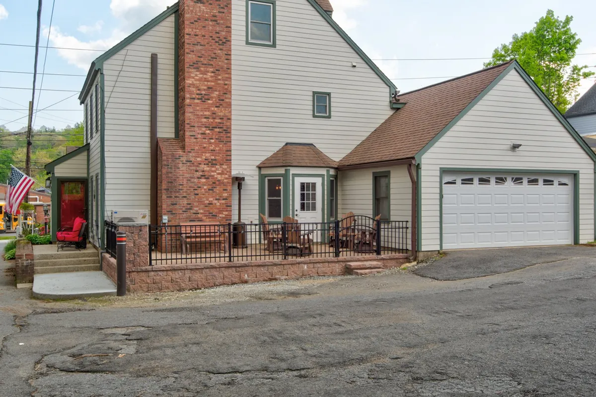 This exterior shot captures the front porch with comfortable seating, a prominent brick chimney, garage door, and the American flag. The porch is bathed in gentle light, perfect for evening conversations or relaxing after a day exploring Blowing Rock.